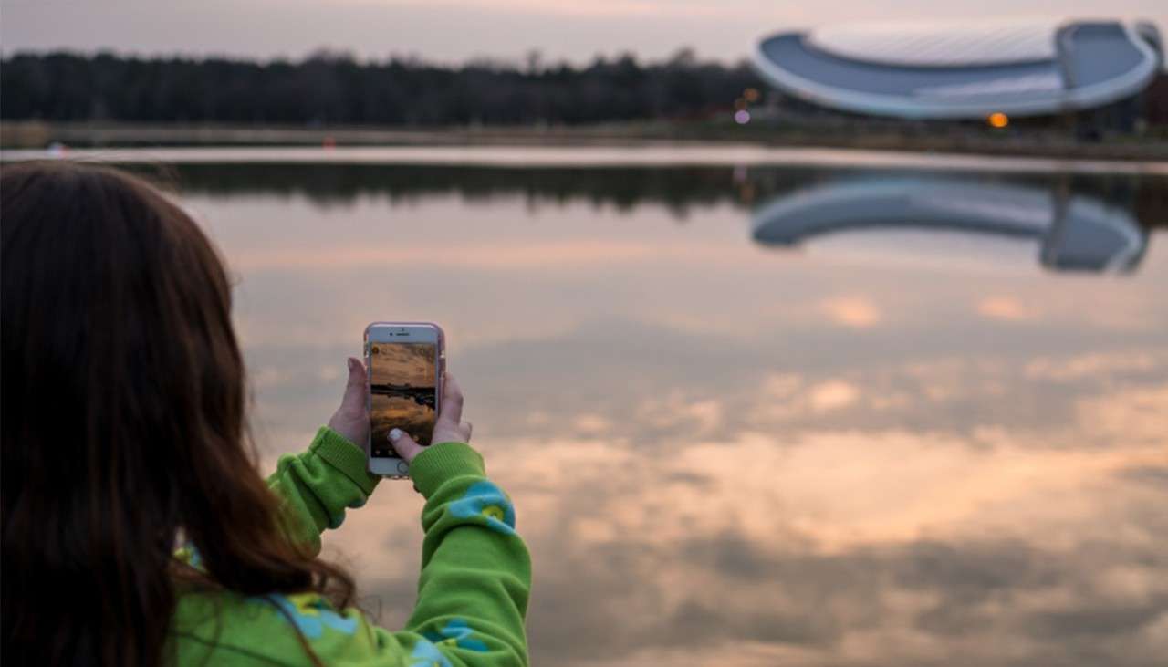 A little girl taking a photo of The Subtropical Swimming Paradise building next to the lake.
