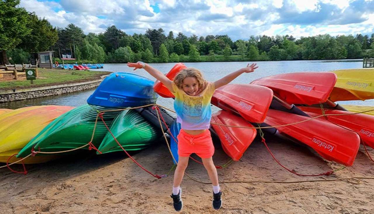 A girl doing a star jump in front of canoes on the beach.