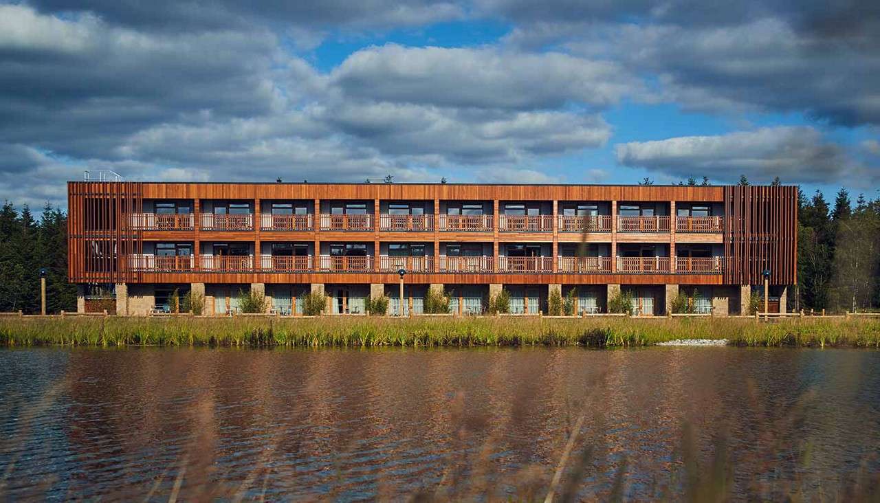 A three-story wooden building faces the camera, displaying rows of balconies, situated beside a calm pond with reeds, backed by dense forest under a cloudy sky.