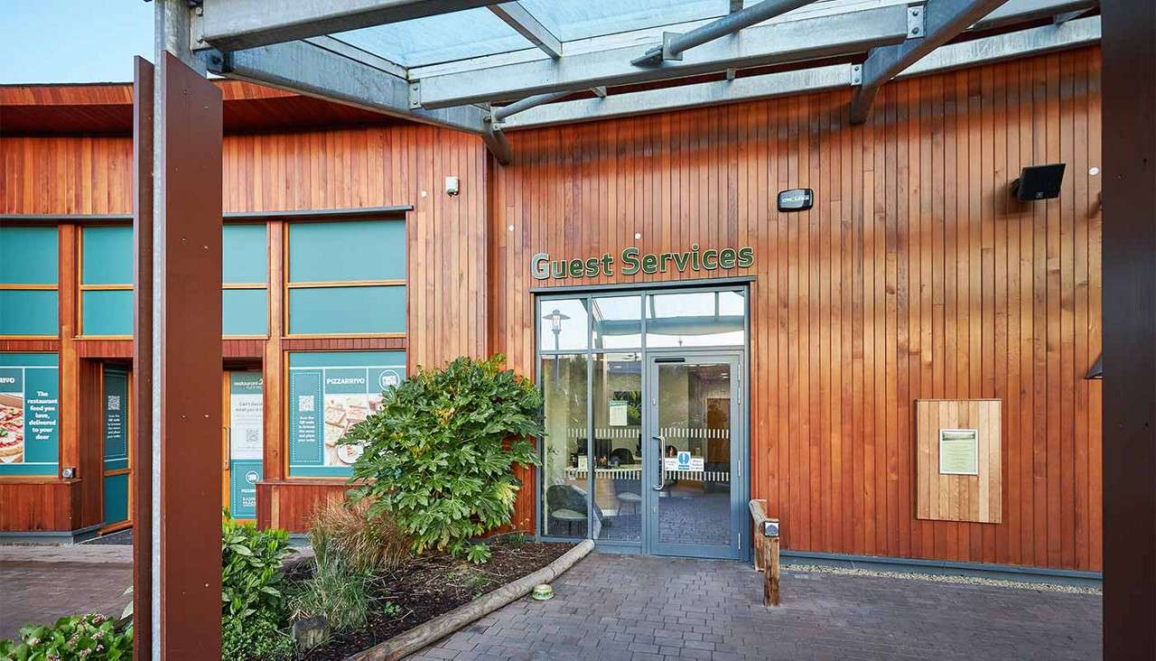 Entrance doors welcome visitors, marked with the sign “Guest Services,” set in a wood‑clad building. Shrubs border a paved walkway under a metal canopy, with nearby windows and posters.