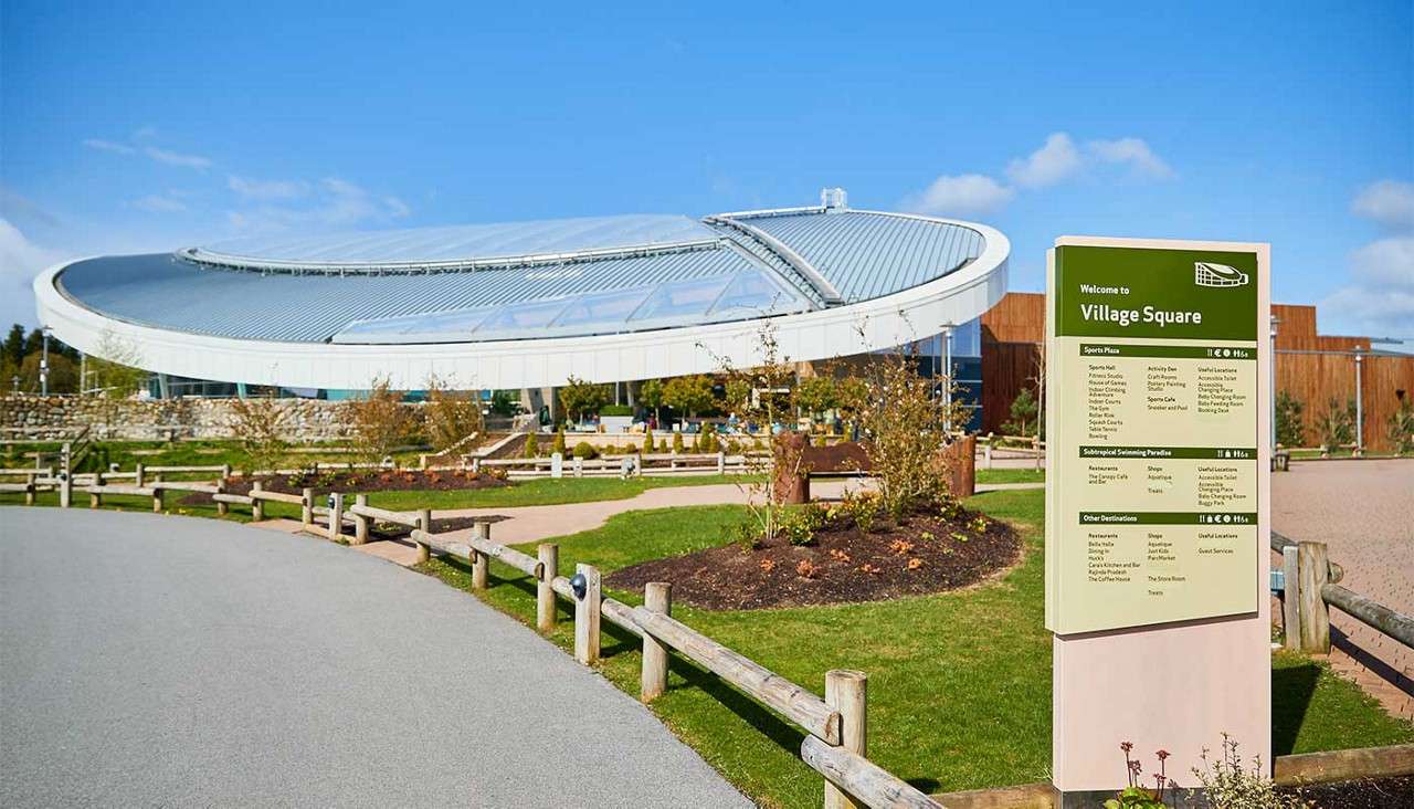 Wayfinding sign lists attractions; a sweeping, glass-roofed circular building sits behind; curved path, garden beds, and wooden rail fences frame the sunny plaza.

Text: “Welcome to Village Square
Sports Plaza
Subtropical Swimming Paradise
Other Destinations”
(Smaller list items are not legible.)