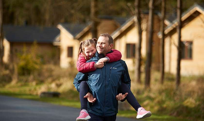 A dad carrying his daughter on his back whilst walking through the forest.