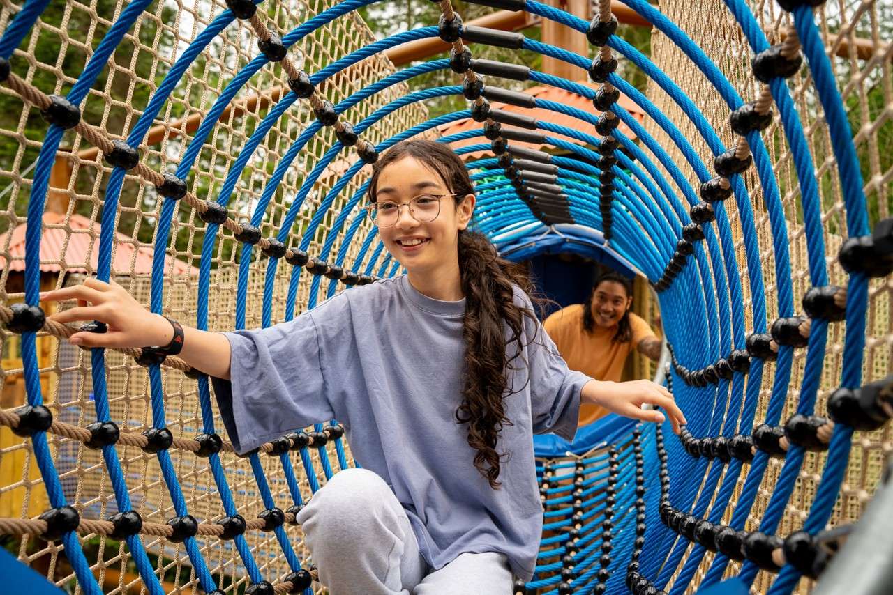 Child smiles and balances while crawling through a blue rope tunnel, gripping net sides. Another person follows behind. Context: outdoor playground with climbing nets and surrounding trees.