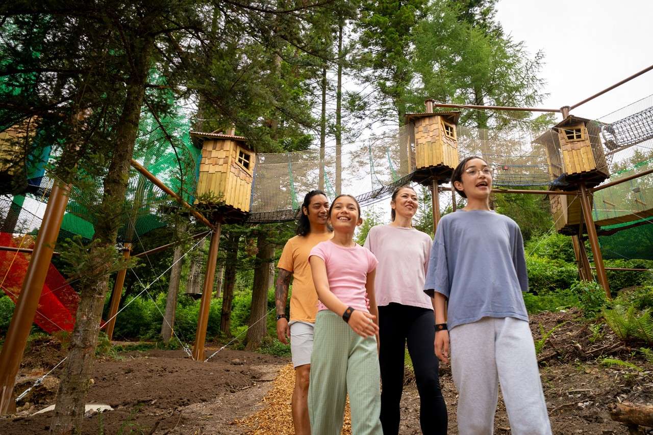 Four people walk and smile beneath elevated rope bridges and small wooden treehouses, surrounded by tall evergreen trees in a forested adventure park.