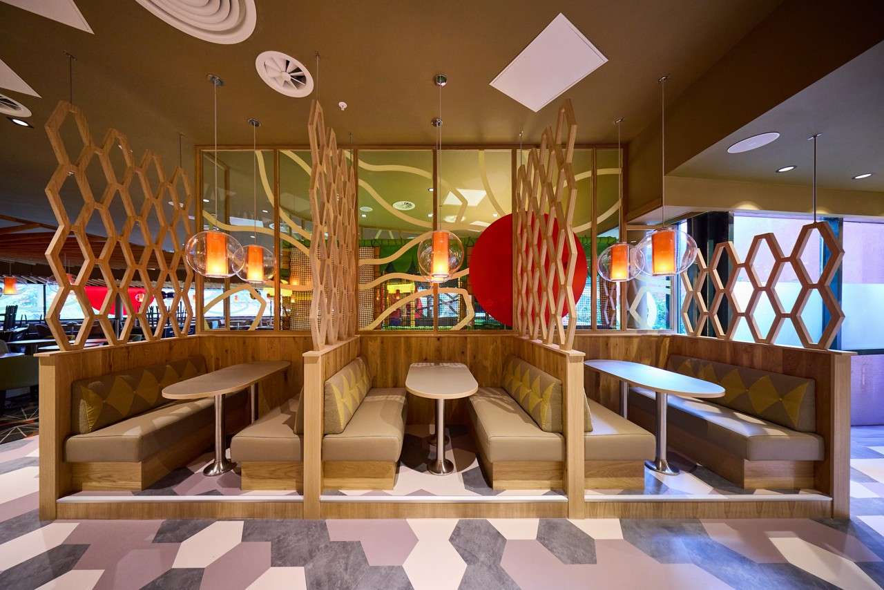 Booth seating invites diners, separated by wooden honeycomb screens, in a modern restaurant; oval tables, tan cushions, pendant lamps, patterned floor, and colorful wall panels fill the surrounding space.
