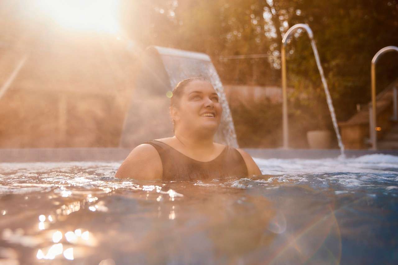 A lady rlaxing in the outdoor pool, the sun rising behind her.