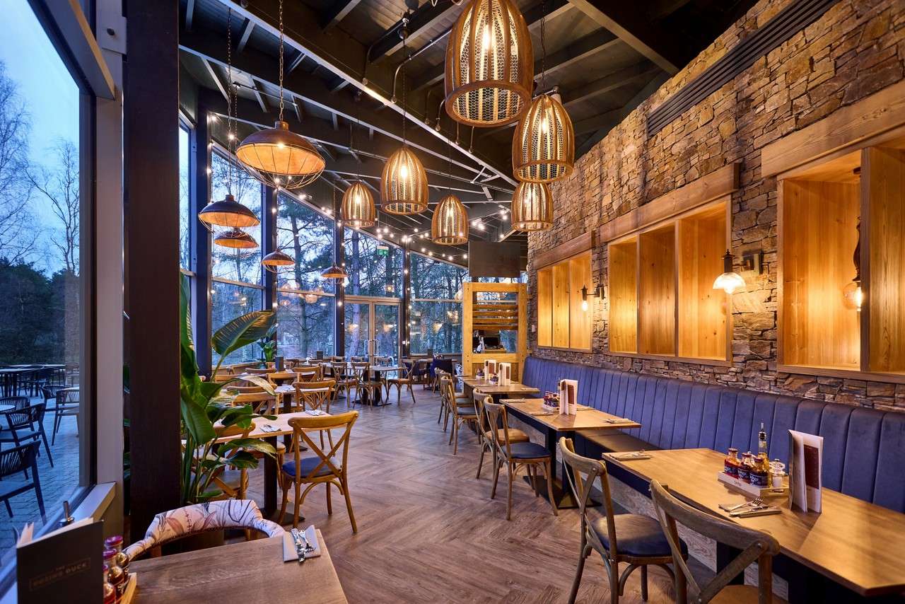 Dining tables await guests, arranged along a blue banquette and wooden chairs, under warm pendant lights, in a rustic-modern restaurant with stone wall and floor-to-ceiling windows overlooking trees at dusk.