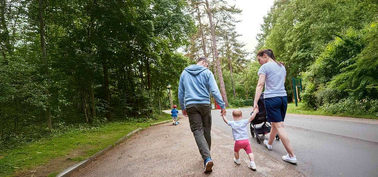 Two adults hold a toddler’s hands while walking, pushing a stroller; another child runs ahead along a paved path through a green, tree-lined park.