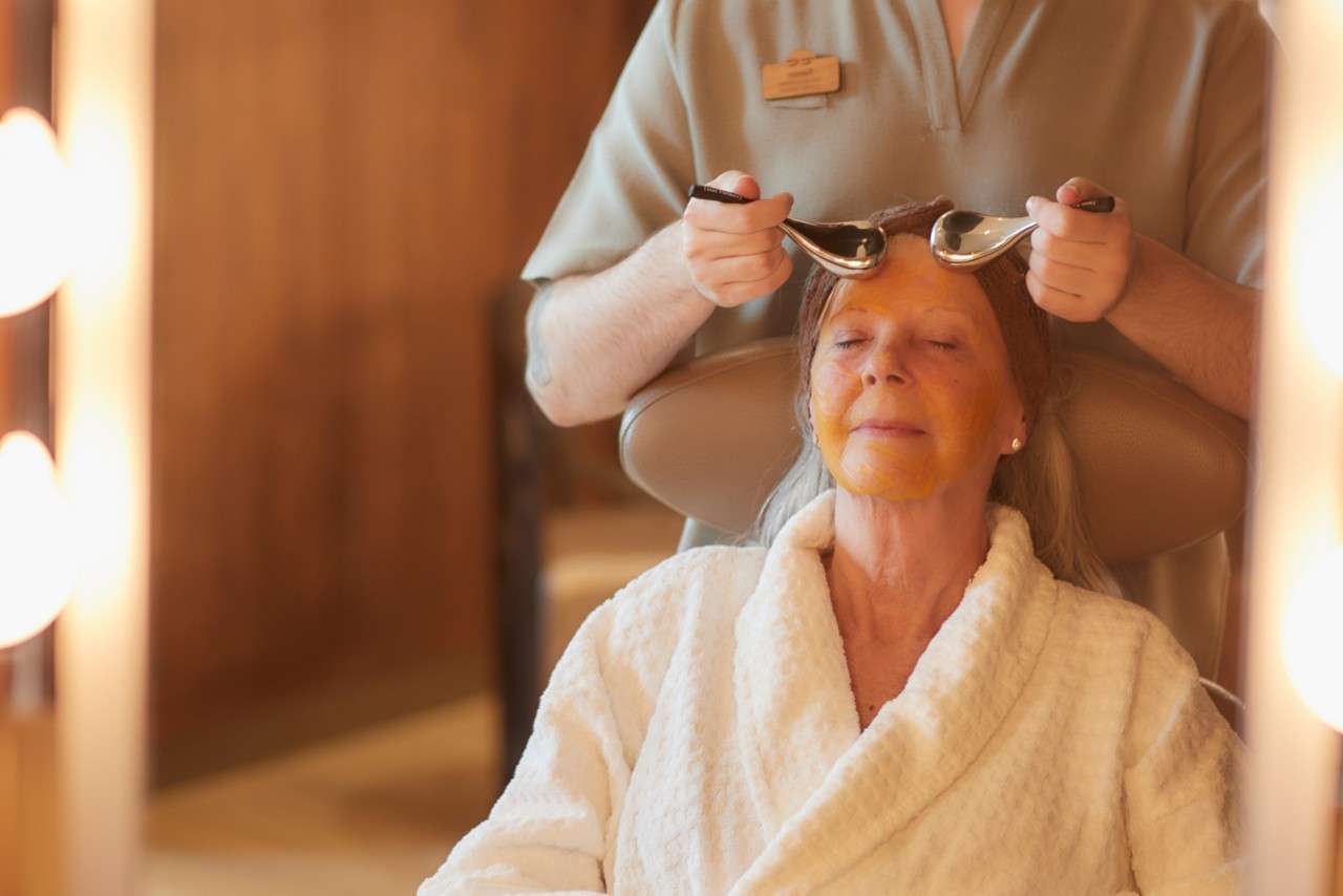 A lady sat relaxing with her eyes closed while a therapist has ice rollers on her face.