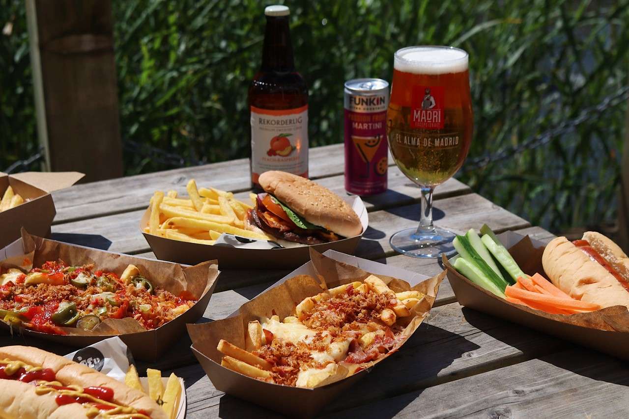 Trays of fries, loaded fries, hot dogs, and a burger sit on a wooden picnic table with beer, cider, and a cocktail can outdoors near greenery. Text: REKORDERLIG; FUNKIN; Passion Fruit; MARTINI; MADRI; EL ALMA DE MADRID.