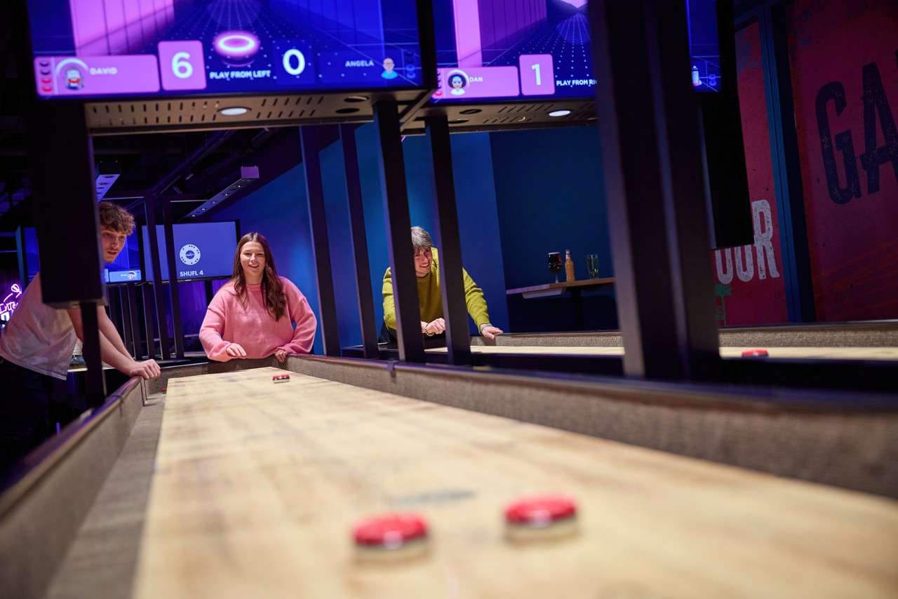Shuffleboard pucks slide down a long wooden table as three people play in a neon-lit arcade bar. Text: DAVID 6, PLAY FROM LEFT, ANGELA 1, DAN, PLAY FROM RIGHT, SHUFL 4.