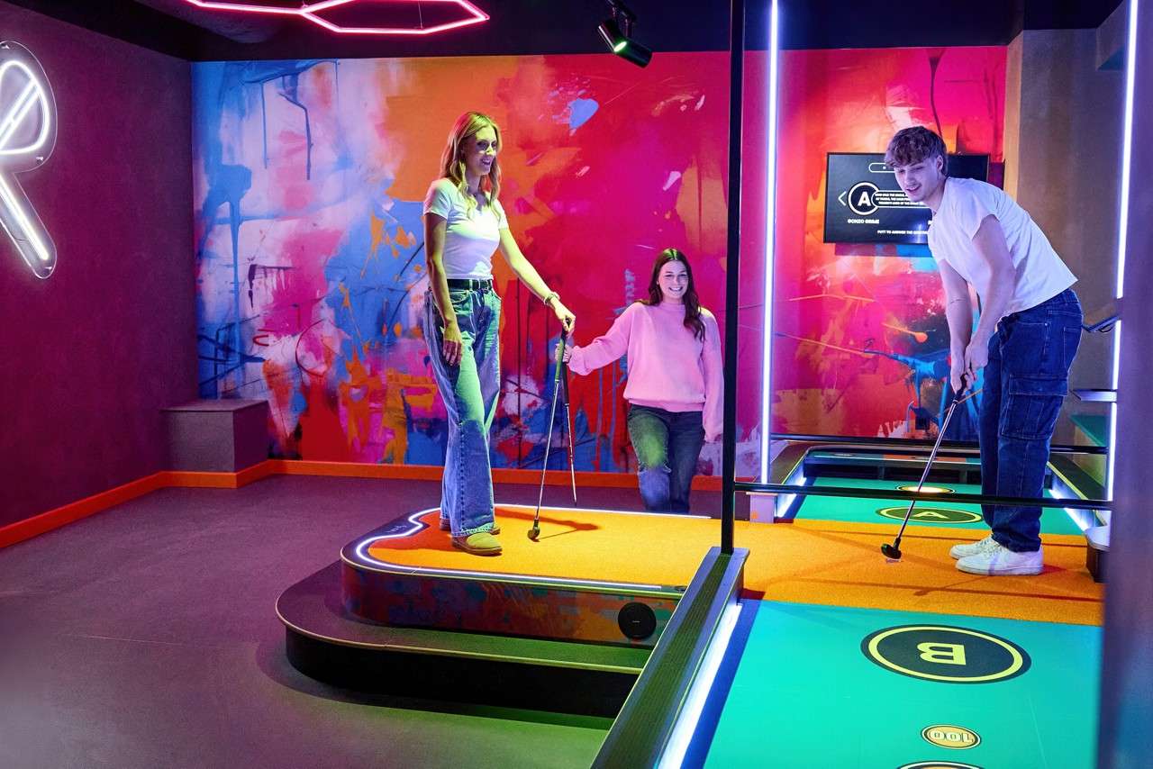 Three people play mini-golf, swinging putters on glowing platforms in a neon-lit room with colorful graffiti walls.
Text: R, A, B, 100.
