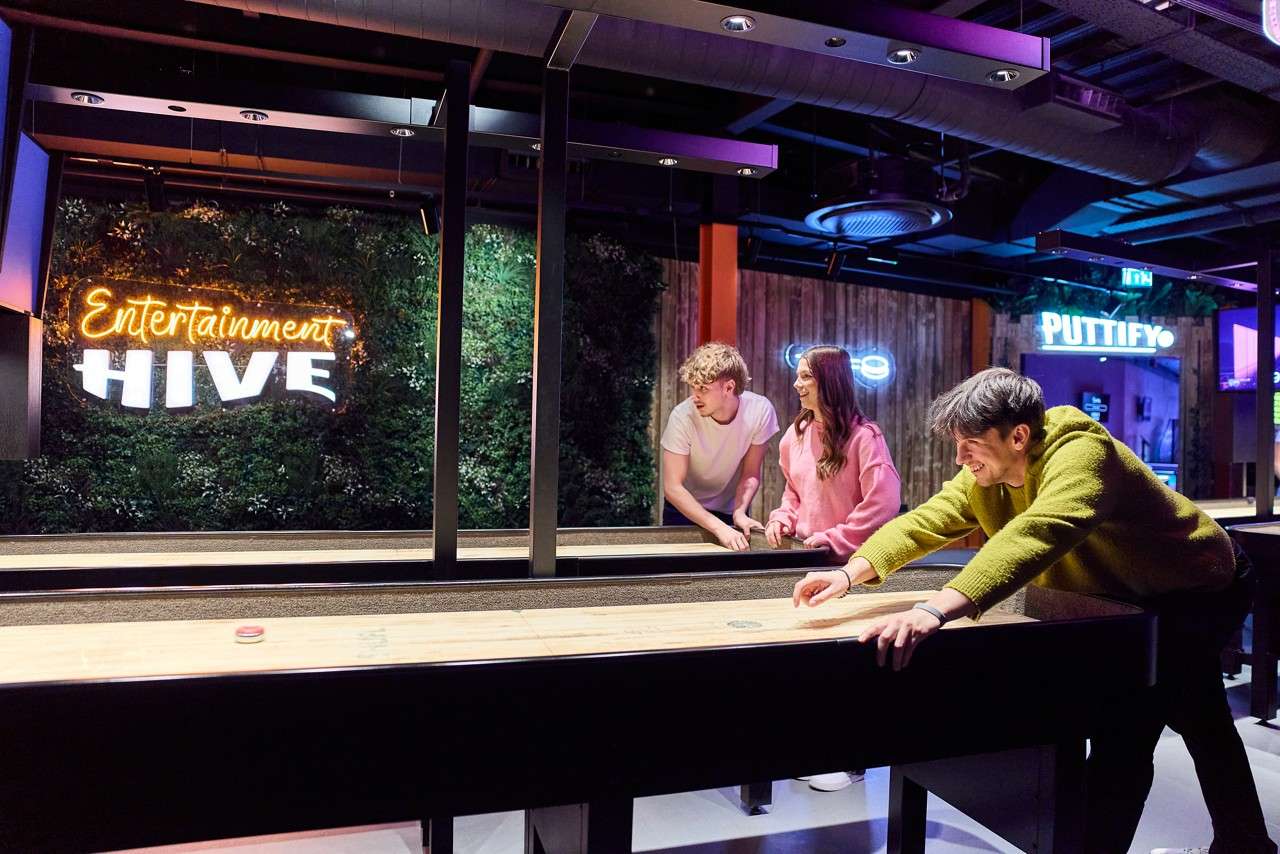 Three people play shuffleboard, sliding a puck down the table, in a neon-lit entertainment venue with greenery wall and signs. Text: "Entertainment HIVE", "PUTTIFY", "EXIT".