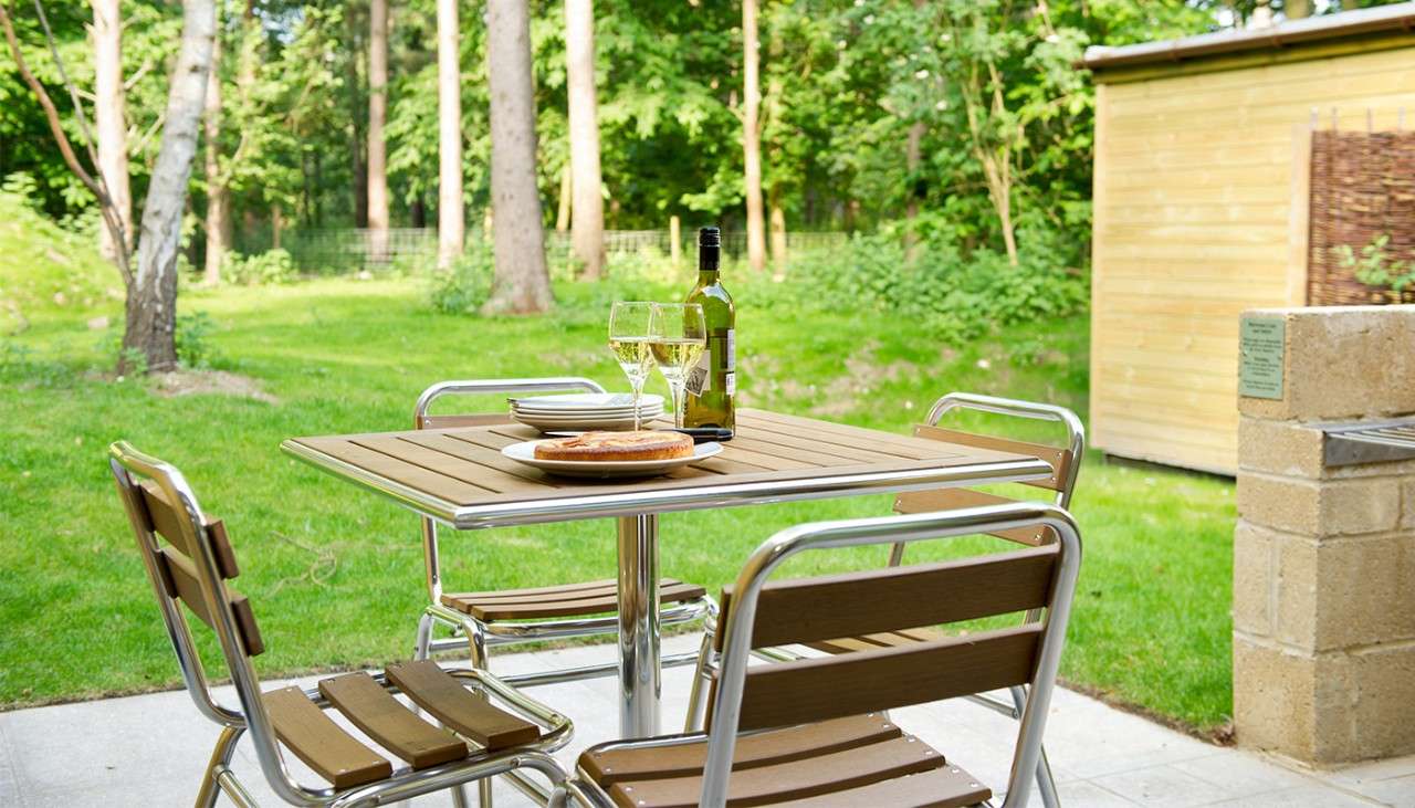 An outdoor patio area showing a table and 4 chairs with a bottle of wine and a plate of food. 