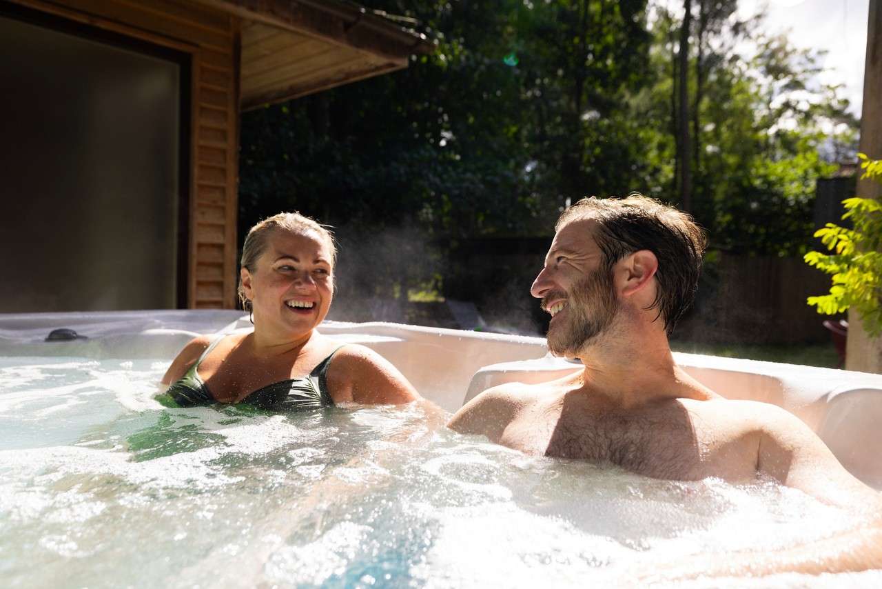 Two adults relax and chat, smiling, while soaking in a steaming outdoor hot tub; sunlight glows on water, with a wooden structure and leafy trees surrounding the yard.