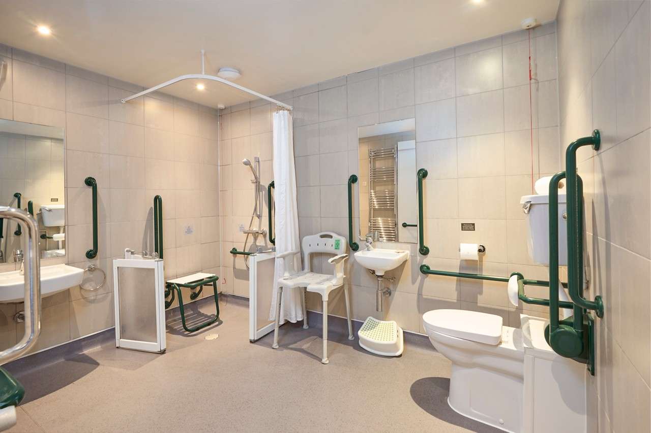 Accessible bathroom—equipped for mobility support. Grab bars line walls; toilet and two sinks with rails; roll-in shower with chair and curtain; fold-down seat; mirrors; bright, tiled, spacious room.