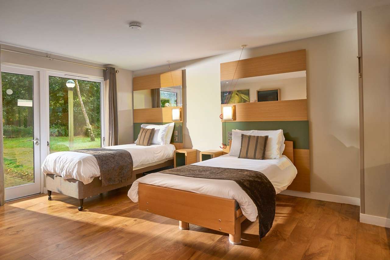 Two single beds sit made with white linens and dark throws, facing bedside lights and mirrors, in a bright room with wood floors and glass doors overlooking a green garden.