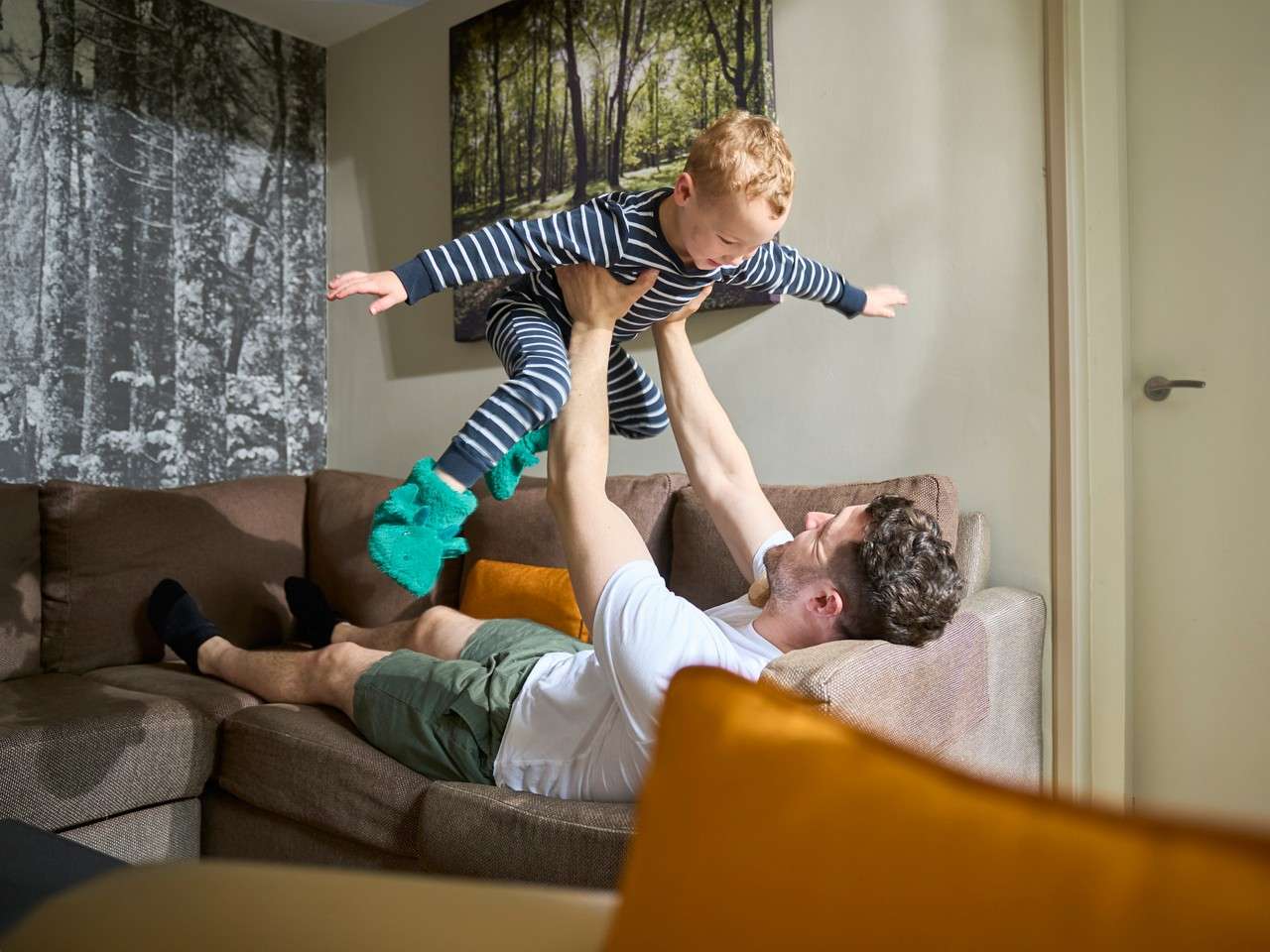 Child in striped pajamas stretches arms as an adult lifts them into the air, both smiling, on a sectional sofa in a living room with forest artwork and orange pillows.