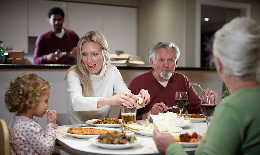 Woman serves flatbread at a round table; child eats, older couple drink wine, and another person stands in the kitchen, in a home dining room with assorted dishes and drinks.