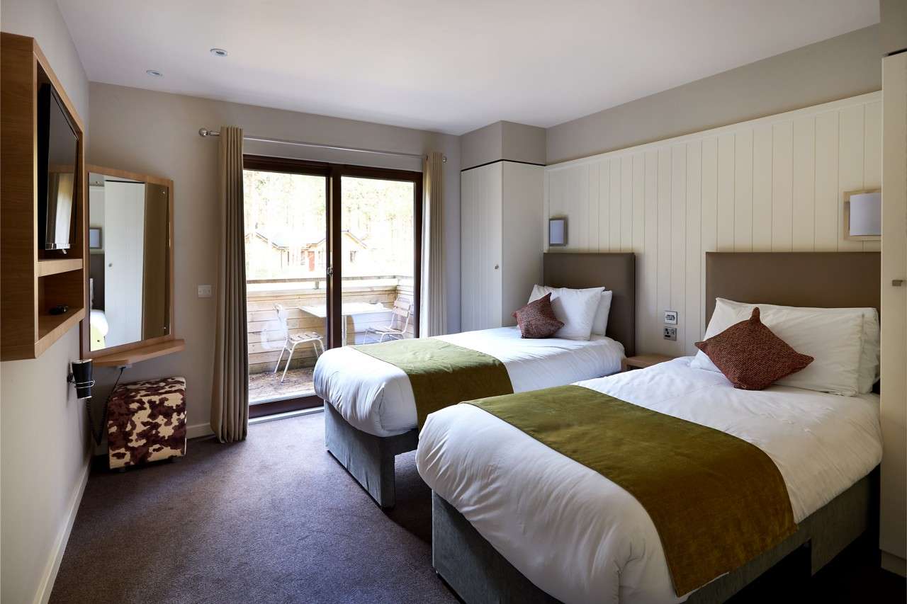 Twin beds stand neatly made with olive runners, facing a wall-mounted TV, in a modern hotel room with sliding glass doors to a balcony, outdoor chairs, mirror, and light decor.