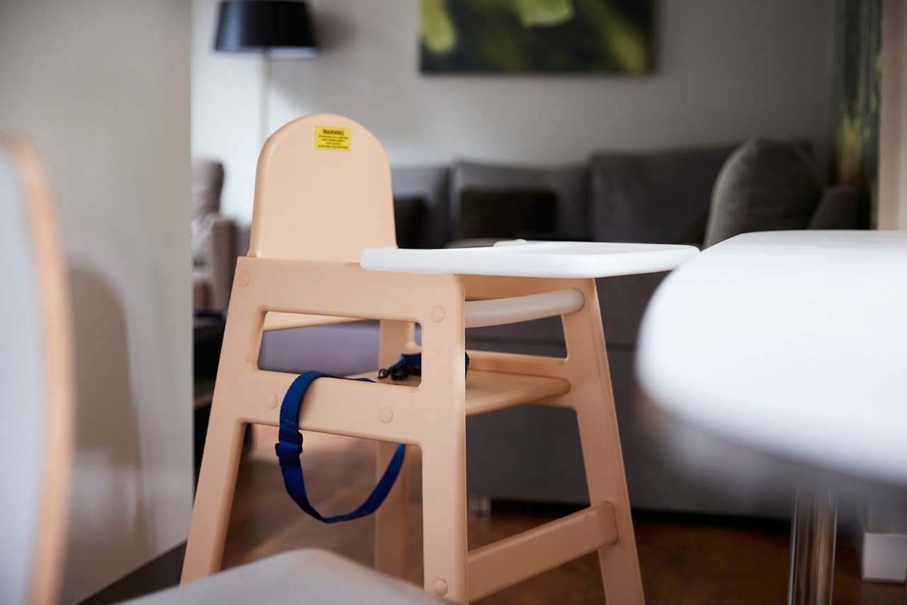 High chair stands empty, blue safety strap hanging; text reads “WARNING.” In a modern living room with gray sofa, floor lamp, wall art, and nearby white tables.