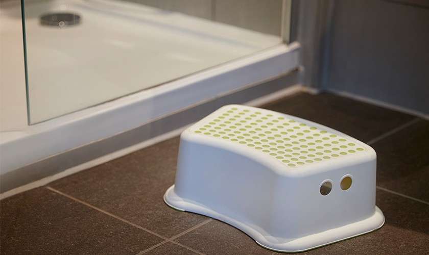 White plastic step stool sits on dark tile, providing elevation; positioned beside a glass-walled shower with raised threshold in a modern bathroom.