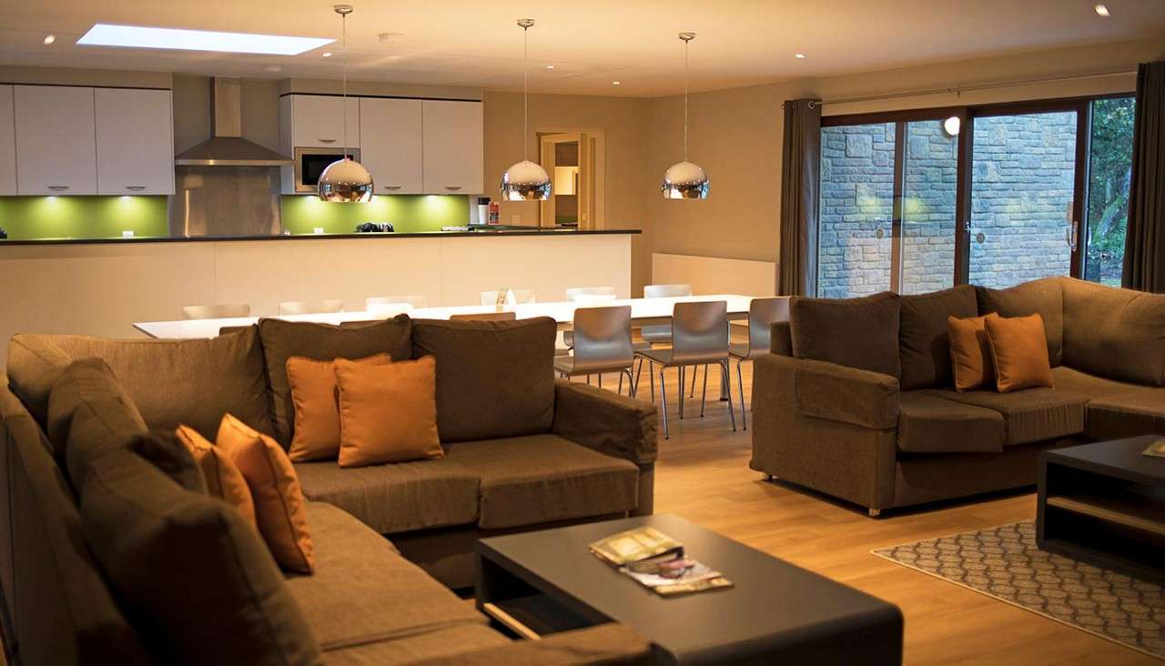 Brown sectional sofas with orange cushions surround low tables; behind them, a long dining table and modern kitchen with pendant lights and green backsplash face large sliding glass doors.