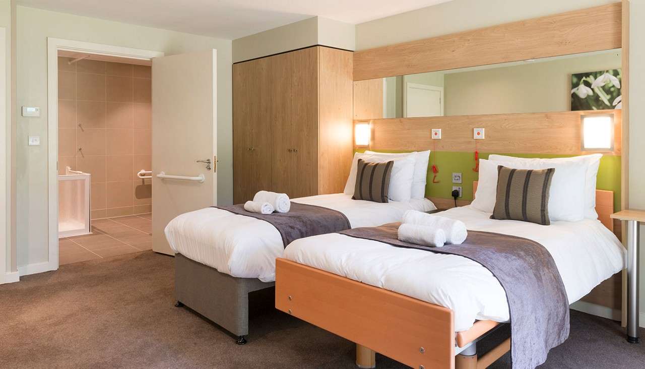 Twin beds sit neatly made with white linens, gray runners, and rolled towels, lit by wall lights; context: an accessible hotel room with wardrobe and open bathroom featuring grab bars.