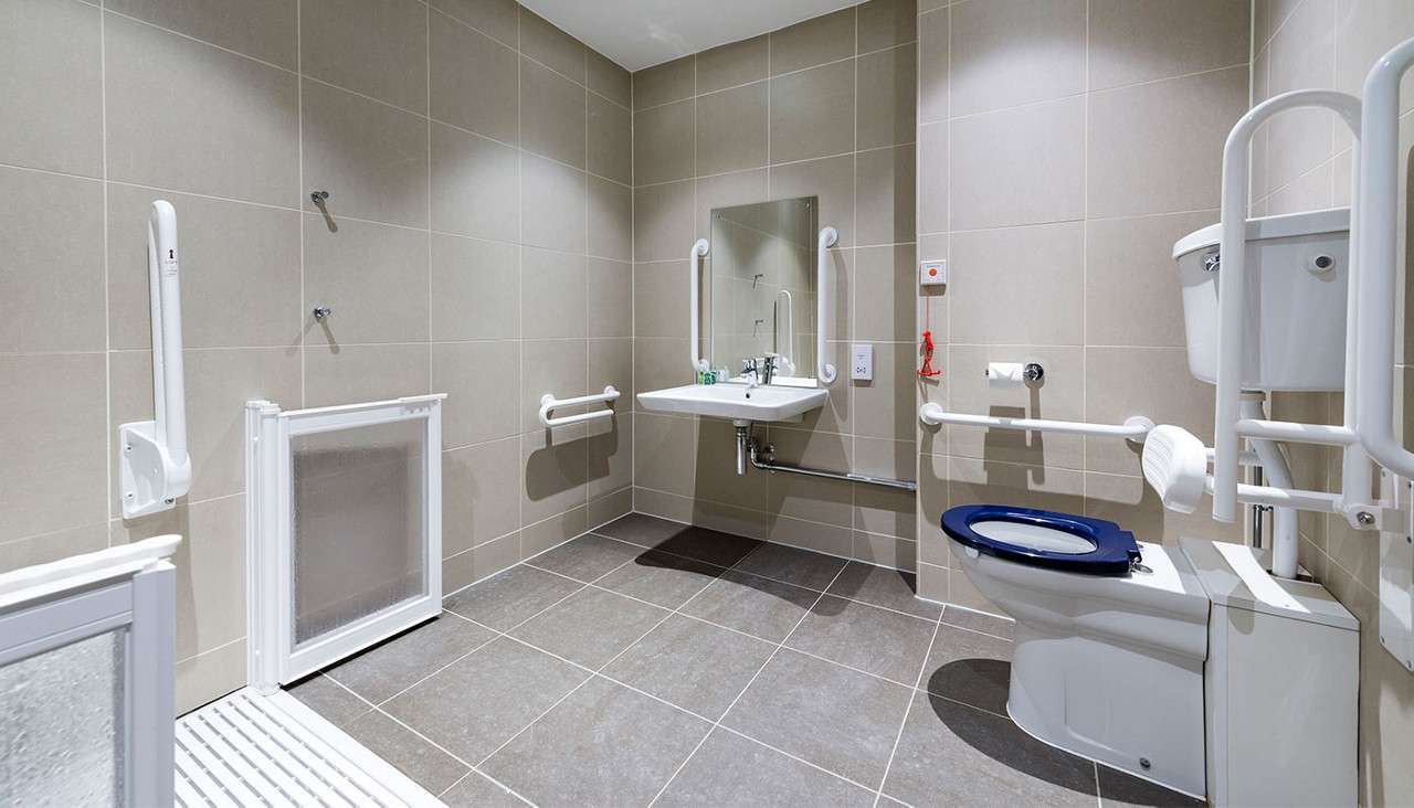 Toilet with blue seat stands between grab bars; sink with mirror and emergency pull cord accompany it; in a spacious tiled accessible bathroom with roll-in shower partition. Text: “1”.