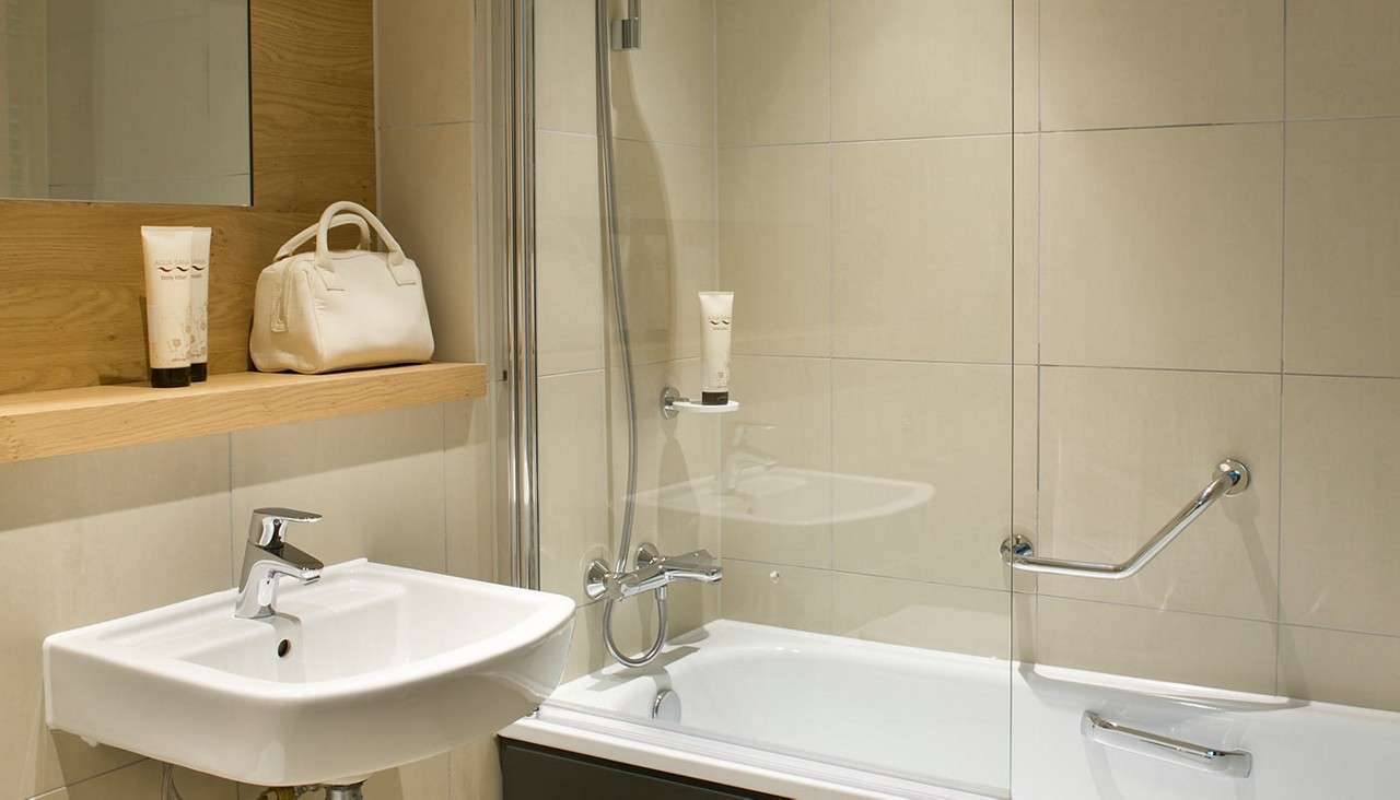 Bathtub and shower stand behind a glass screen, with chrome grab bars attached; nearby, a white sink and wooden shelf hold toiletries and a white bag in a beige-tiled bathroom.