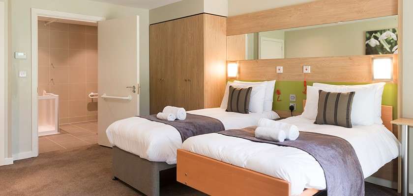 Twin beds sit neatly made with gray runners and rolled towels, facing a wide mirror and wall lamps; nearby a wooden wardrobe and open doorway reveal a tiled accessible bathroom.