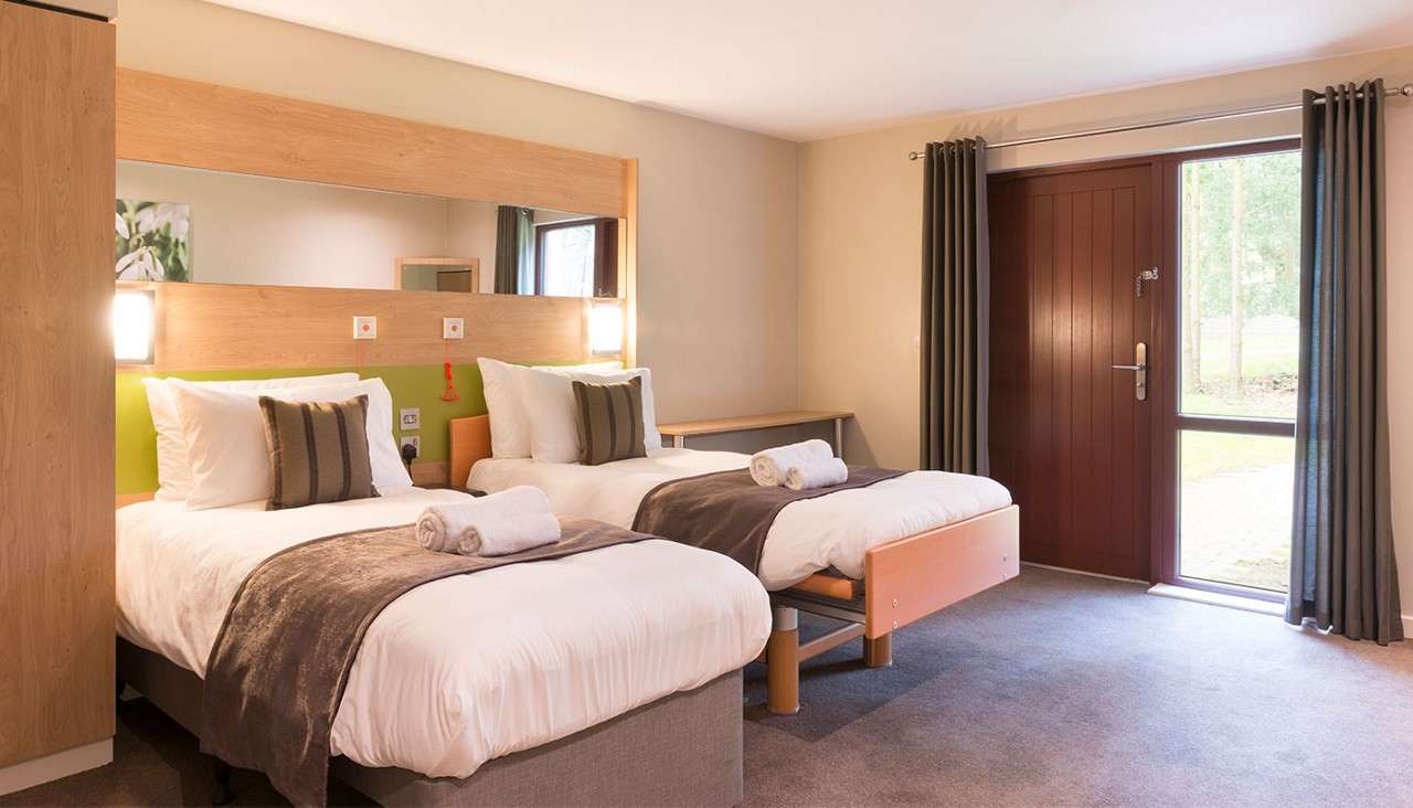 Two single beds with white linens and rolled towels sit side by side; a large mirror and wall lights above. Context: simple room with desk, wooden door, and curtained window.