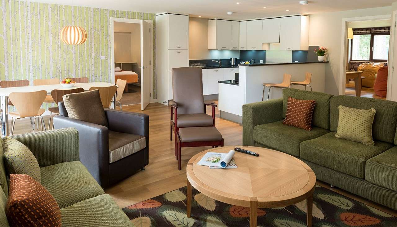 Green sofa and armchairs surround a round wooden coffee table holding magazines and a remote; behind, a modern kitchen with breakfast bar and dining table, with open doorways leading to bedrooms.