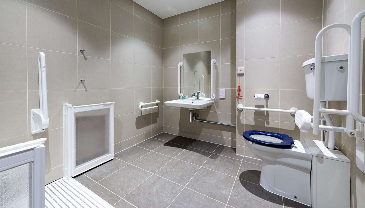 Accessible bathroom supports users: grab bars frame sink and toilet, emergency pull cord hangs, open shower area with folding screen. Context: spacious, beige-tiled room with bright lighting and floor-level drainage.