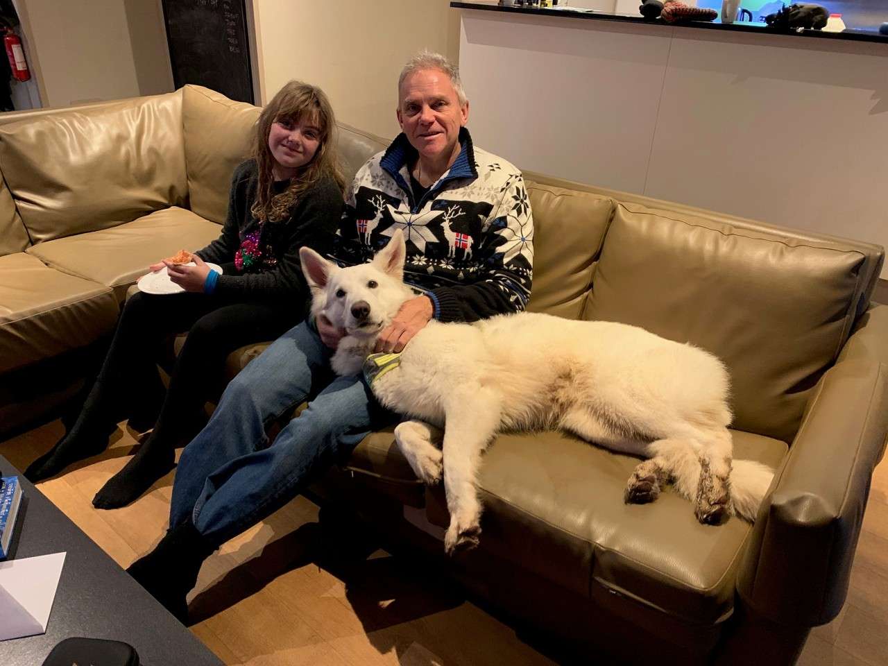 White dog lounges across a leather sofa, head on a man’s lap, muddy paws visible; a man and girl sit together, the girl eating pizza, in a cozy living room.