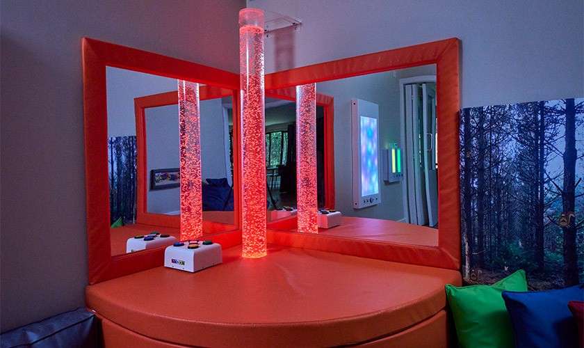 Two vertical bubble tubes glow red, bubbling upward; they stand on a padded corner table flanked by mirrors, with small controllers and wall-mounted light panels in a sensory room.