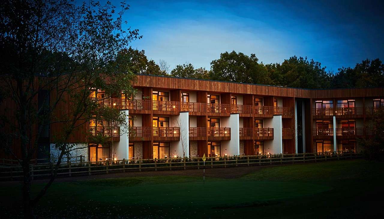 Modern three-story building glows with warm balcony lights, facing a fenced lawn and small golf green. Surrounded by trees at dusk, the structure curves gently beneath a darkening sky.
