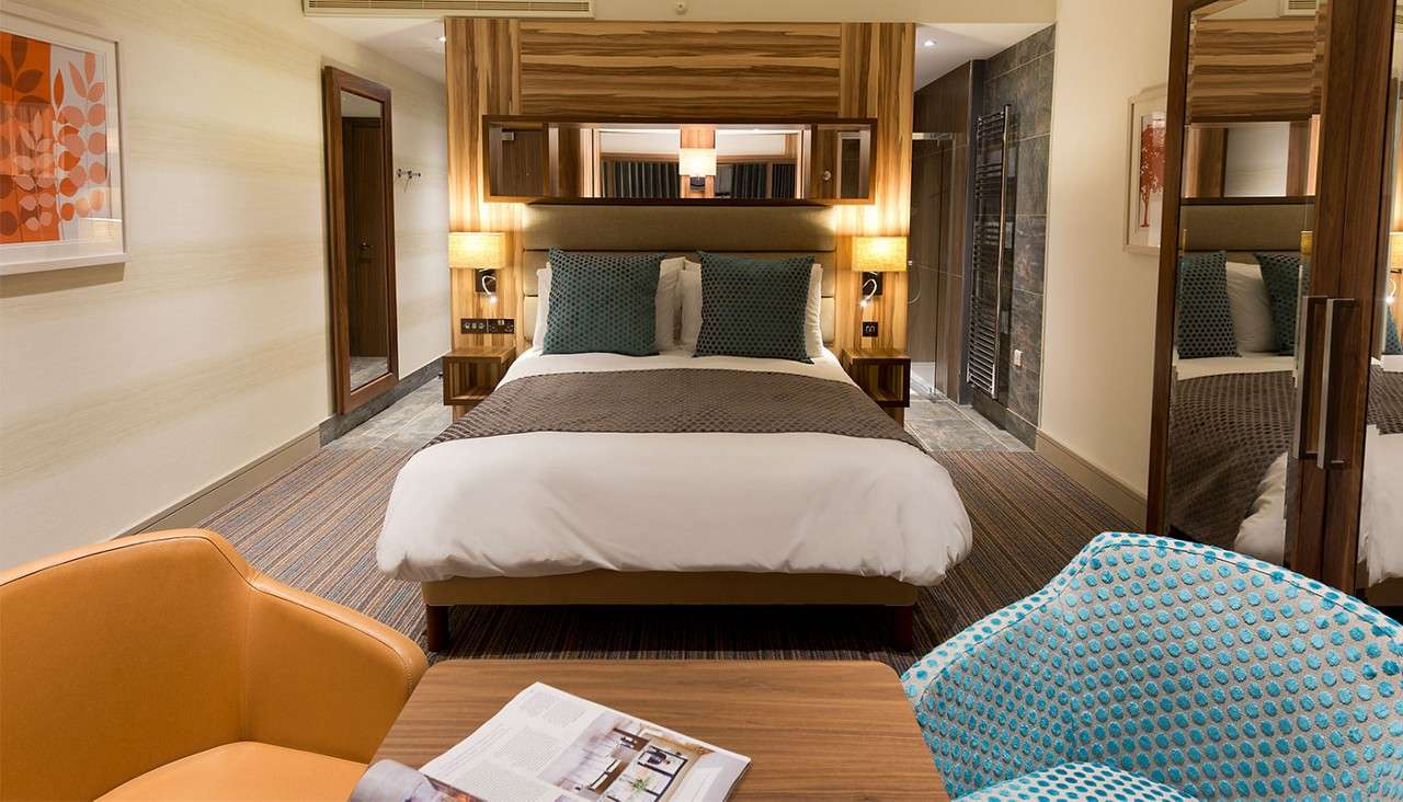 Bed anchors the room, neatly made with two teal pillows, flanked by lamps; a hotel room with carpet, wooden headboard, chairs, table with open magazine, and a doorway to bathroom.
