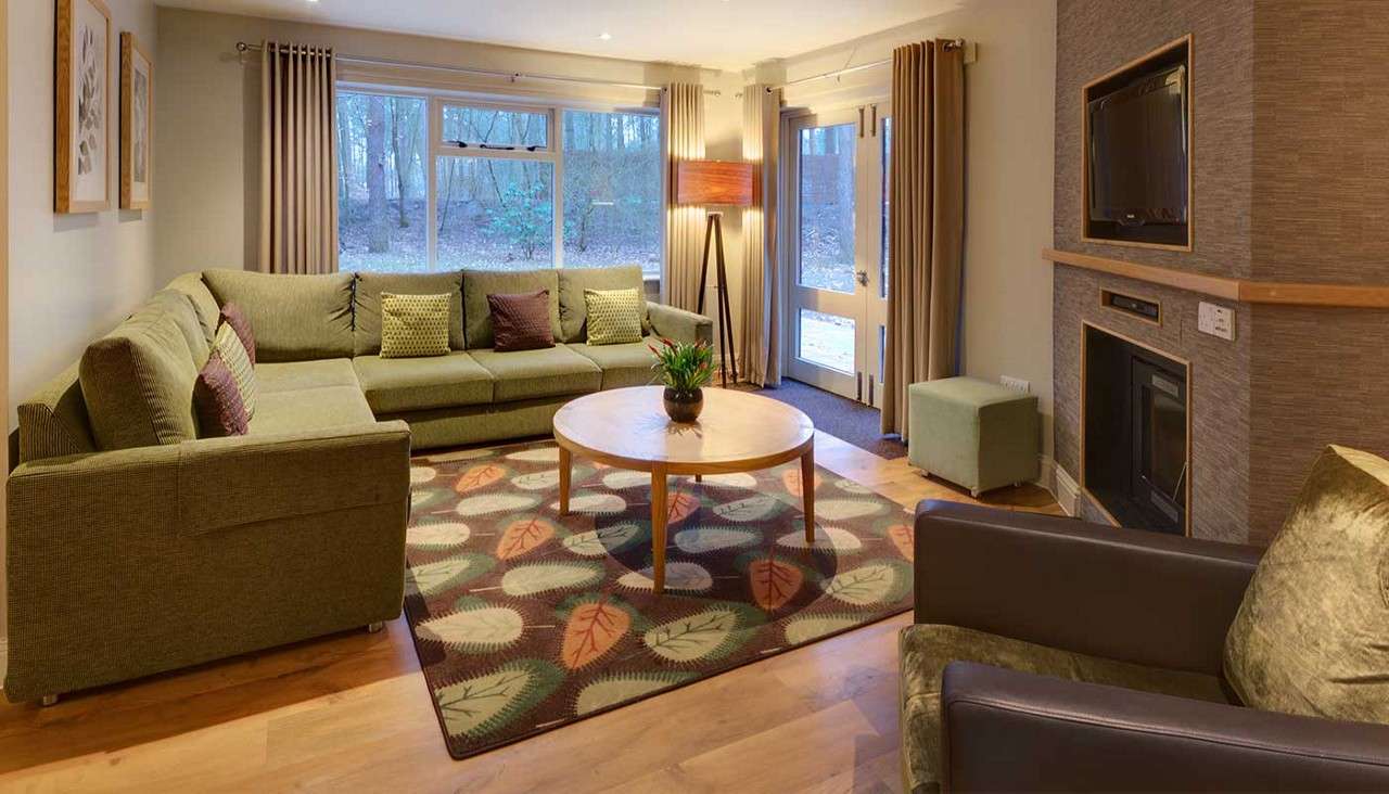 Green sectional sofa anchors seating, arranged around a round wooden coffee table, in a living room with patterned rug, floor lamp, fireplace with TV above, windows and door overlooking woods.