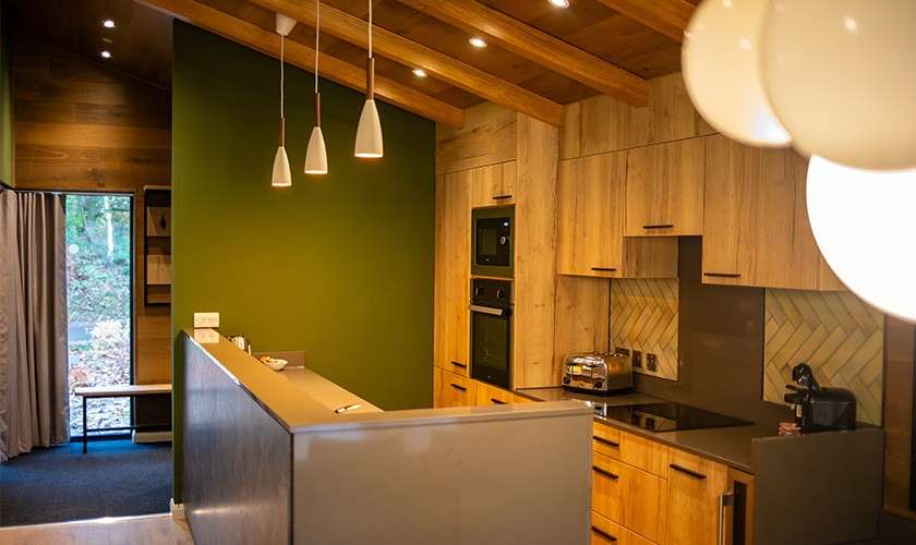 Kitchen gleams under pendant lights, displaying wooden cabinets, built-in double oven, induction cooktop, toaster, and coffee maker, within a cozy green-walled room with timber ceiling beams and window to trees.