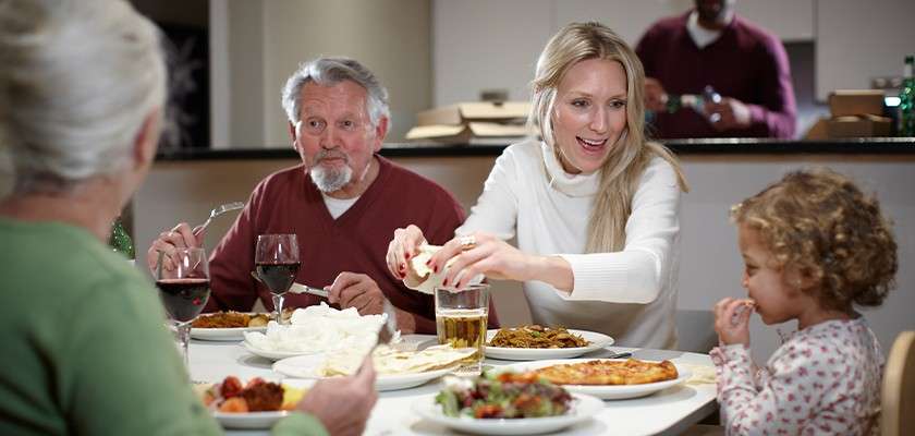 Family diners share a meal; adults serve food while a child eats, at a white table with pizza, salad, wine, and beer, in a home kitchen background.