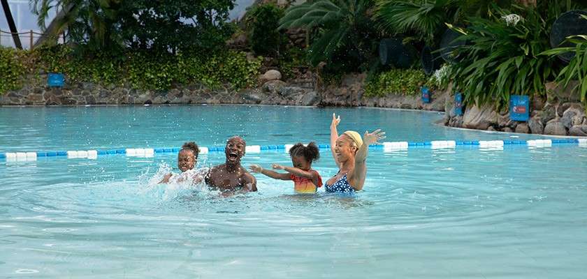 Family of four splashes and laughs, waist-deep in a swimming pool. Tropical resort surrounds them with stone edging, lush plants, and blue-white lane markers floating on the calm water.