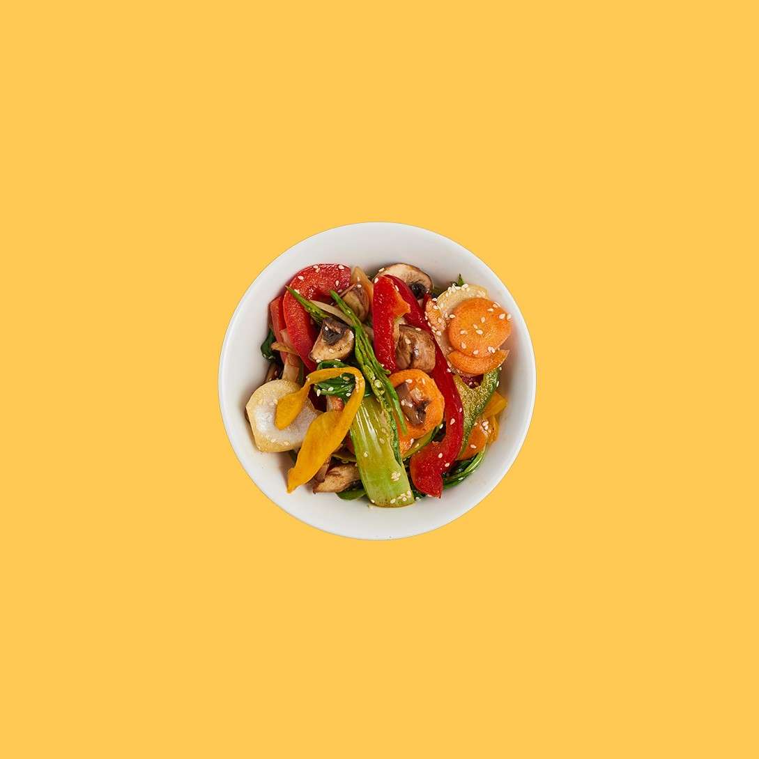 A white bowl holds stir-fried vegetables—bell peppers, carrots, mushrooms, bok choy—glistening with sauce, sprinkled with sesame seeds, centered against a plain mustard-yellow background.