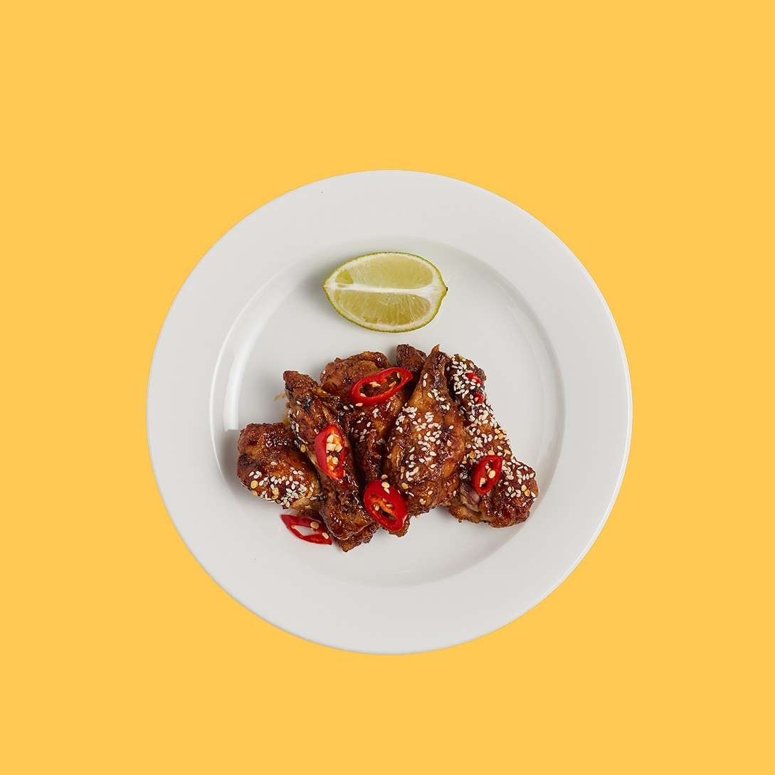 Chicken wings—glazed, sprinkled with sesame, topped with red chili slices—rest on a white plate with a lime wedge, against a bright yellow background.