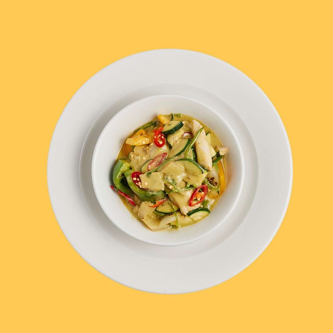 Vegetable curry sits in a white bowl, showcasing zucchini slices, bamboo shoots, peppers, and red chili in creamy green sauce; placed on nested white plates against a solid yellow background.