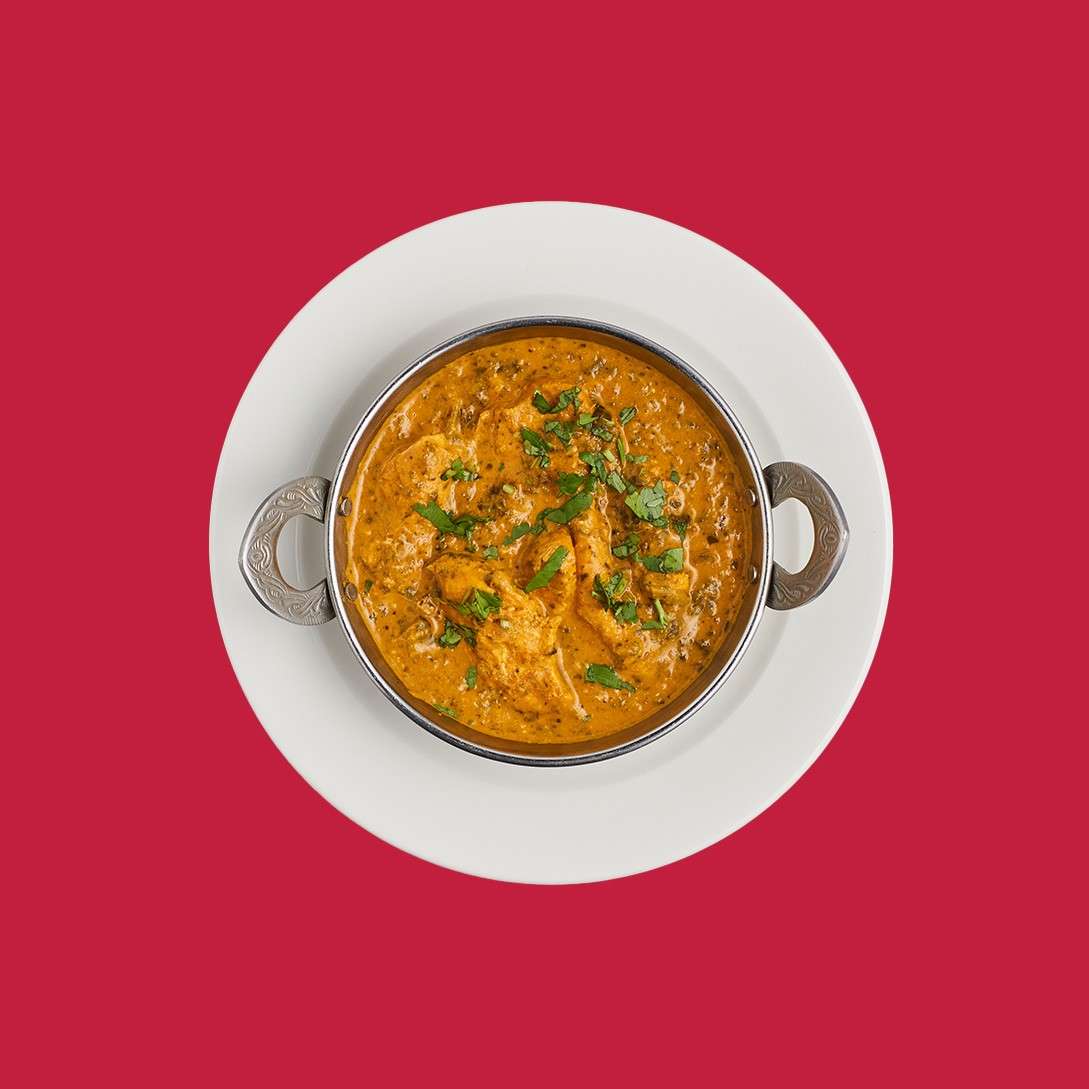 Silver-handled bowl of orange curry rests on a white plate, garnished with chopped green herbs, thick sauce with chunks, centered against a solid red background.