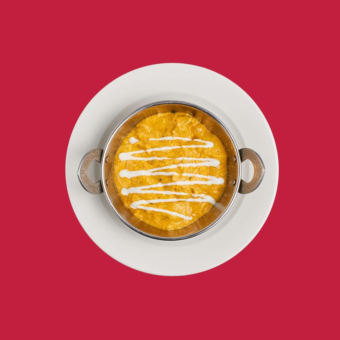 Bowl of orange curry drizzled with white sauce sits centered on a white plate against a solid red background.