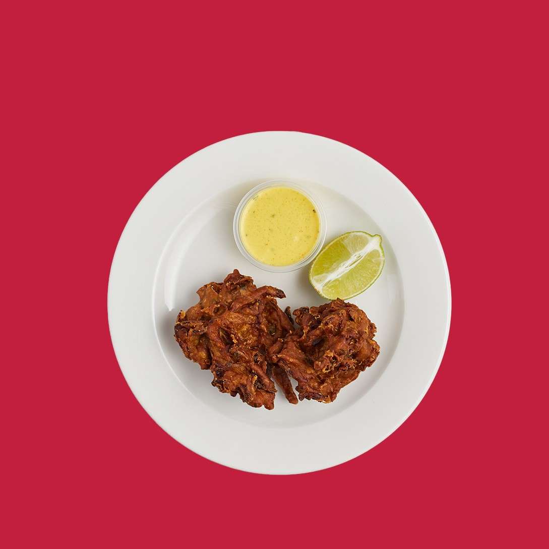 Fried fritters sit beside a small cup of yellow dipping sauce and a lime wedge, served on a white plate centered against a solid red background.