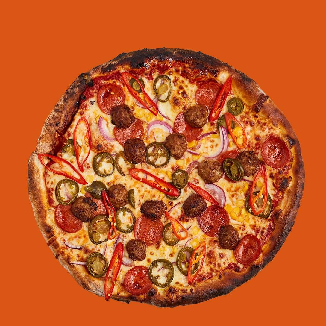 Pizza sits flat, topped with pepperoni, meatballs, jalapeño slices, red chili strips, red onion, and melted cheese; browned crust. Set against a solid orange background.