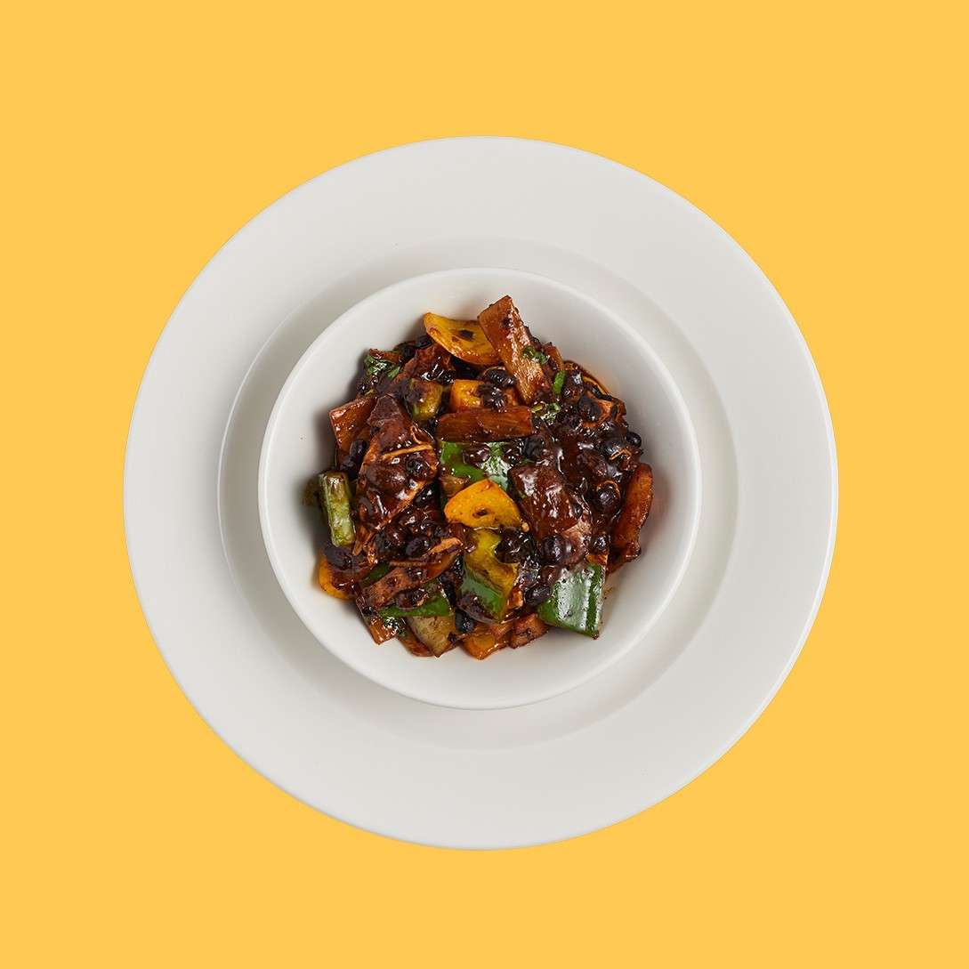 A white bowl of dark, saucy stir-fry rests centered, with peppers, onions, and chunks of protein, set on a white plate against a solid yellow backdrop.